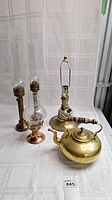 Photo of the brass teapot, lamp base, and three candle holders arranged on a white cloth background.