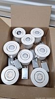 Top-down view of eight white recessed ceiling lights in an open cardboard box showing the 7-LED lamp cluster and metal housings.