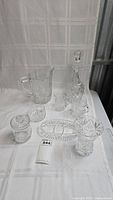 Full view of all crystal glassware items arranged on a white cloth background showing lidded bowl, creamer, sugar bowl, water pitcher, decanter, and stopper.