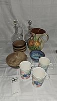 Full view of all items including two glass decanters with stoppers, ceramic pitcher with floral design, stoneware bowls and three fruit patterned tea mugs.