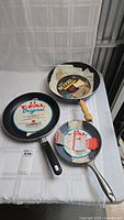 Three frying pans positioned on white surface: one Revere with wooden handle, two T-fal Original with black handles and original labels.