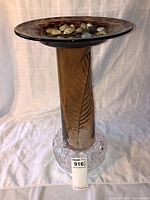Full view of the handmade glass birdbath or plant stand, showing the shallow top with stones, tall brown stem with leaf design, and clear textured base.