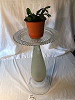 Full view of tall handmade glass plant stand with a small potted plant placed on top. Clear ribbed glass base and platform, frosted central column with a weathered appearance.