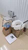 Photo showing the foot bath, bathtub/shower seat with gray backrest and metal legs, raised toilet seat with black support handles, a small box with miscellaneous health items, and a white digital scale on the floor.