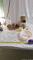 Lot overview showing all items on white cloth including glass plate, casserole dish, fruit-shaped ceramics, serving tray and Medalta pot