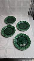 Four green majolica plates laid flat showing front with leaf pattern and textured border detail.