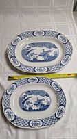 Two Old Chelsea oval porcelain platters in blue and white, side by side on a white tablecloth. A measuring tape shows approximate size of each platter.