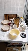 Full view of lot items on white cloth. Includes Pyrex bowl, ceramic bowls, candle holders, coffee pot, mugs, decorative brass item, and other kitchenware.
