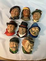 Eight Bossoms chalkware figural wall plaques laid out on a white surface showing a variety of men’s facial expressions and headwear including a Native American, fisherman, elderly men, and a man with top hat.