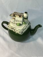 Photo of the dark green ceramic teapot with detailed lid featuring a picnic stove, bottle, and fruit