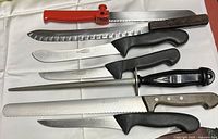 Top view showing seven knives and one honing steel arranged horizontally on a white surface, including serrated bread knives and butcher knives with various shaped handles and blades.