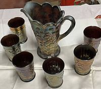 Set of one carnival glass pitcher and six matching drinking glasses arranged on white cloth. Glasses have iridescent finish and floral embossed design.
