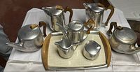 Full set of Mid-Century Modern Piquot Ware tea and coffee set displayed on white cloth including two teapots, coffee pot, creamer, sugar bowl all with wooden handles and a serving tray with wooden handles and woven inset.