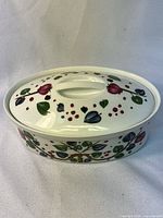 Oval white ceramic casserole dish with hand-painted red, purple-blue, and green floral and heart decoration, with matching lid and integrated handle.