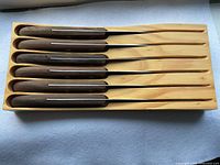 Six steak knives neatly arranged in a wooden tray, showing dark wood handles and stainless steel blades.