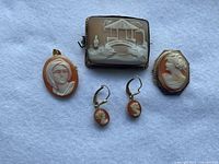 Front view of five cameo jewelry pieces on white fabric including three brooches and a pair of earrings showing relief white cameo designs on orange and brown backgrounds