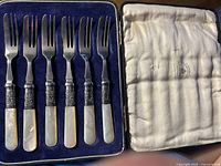 Set of six Mappin Webb pastry forks with mother of pearl handles neatly arranged in original blue velvet-lined box with white satin interior lid showing Mappin Webb branding.