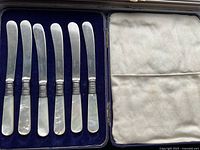 Full set of six dessert knives with mother of pearl handles displayed in blue velvet case with white satin lid.