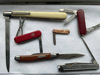 Seven pocket knives on a white cloth background showing handles and some open blades, including a red Victorinox multi-tool.