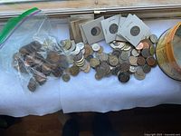 Photo showing a variety of loose coins and several in protective holders, along with coins in a plastic zip bag, all displayed on a white background near a window.