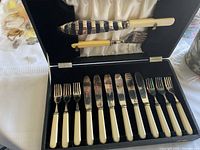 Full view of the fish cutlery set in its original black lined case with satin interior showing fish knife, fish fork and 12 pieces of matching cutlery with cream handles.