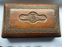 Close-up photo of the wooden box lid, showing detailed hand-carved RAF emblem featuring wings and crown inside an ornate floral border.