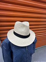 Front angle view of natural paper straw hat with black band against orange metal background.