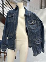 Front and side views of blue denim-style stretch jacket with flap pockets and button closures, displayed on mannequin.