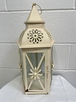 Front and side view of white metal decorative lantern candlestick with glass panels and floral design. Shows the overall shape, peaked roof, and floral cut-out details on metal.