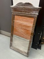 Full view of the ornate wooden mirror wrapped in protective plastic, showing its shape and size leaning against a wall.