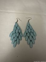 Front view of turquoise fine bead drop earrings arranged flat on a beige fabric surface, showing overlapping glossy elongated oval beads on metal hooks.