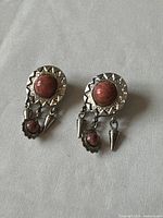 Pair of vintage earrings shown side by side on grey fabric background, highlighting large pink cabochon stones set in a silver-tone geometric base with dangling cone and smaller pink stone elements.
