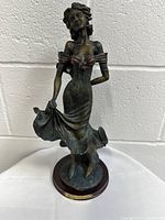 Front view of the statuette showing the woman in a flowing dress posed standing on a round wooden base.