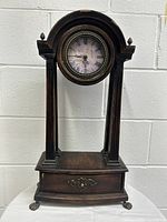 Full view of vintage wooden clock showing round clock face, wooden columns, and drawer base with metal feet
