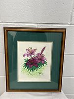 Full view of framed watercolor painting showing purple lilies with green leaves against white background, wood frame and green mat.
