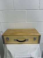 Front view of wooden carrying case placed on white cloth surface showing natural wood finish, black handle and metal latches