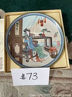 Full view of the Asian decorative plate inside its box, showing the entire painted scene and border.