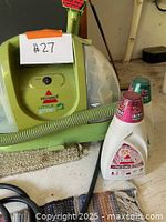 Photo of green and white Bissell Little Green carpet cleaner with hose, two bottles of cleaning solution, and a woven rug underneath.