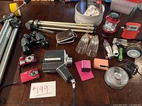 Photo showing assortment of cameras, binoculars, tripods, lanterns, flashlights, small electronic devices and accessories arranged on a table.