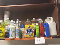 Shelf with a variety of cleaning products including spray bottles, liquid cleaners, soap pads, and sponges.