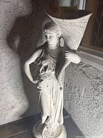 Front and side views of the white ceramic garden statue of a lady holding a cornucopia horn, placed on a tiled surface near a textured wall and a window.