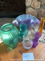 Eight assorted glass vases and one large decorative glass bowl arranged on a wooden surface with stone background, showing colors and shapes including green, purple, amber, and clear glass.
