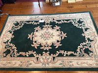 Full view of the green carpet with cream and pink floral details and border lying flat on hardwood floor.