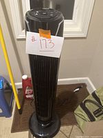 Full view of the black oscillating tower fan standing upright with an attached numbered tag