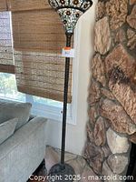 Full view of the Tiffany style floor lamp showing stained glass floral shade, twisted metal column, and ornate base near a stone wall and window.