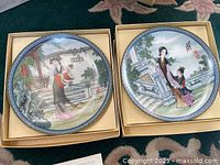 Two decorative Asian porcelain plates in original beige cardboard boxes on floral carpet