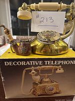 Photo showing gold-tone decorative rotary telephone with cream handle and circular decorative dial on top of the original box labeled 'Decorative Telephone'.