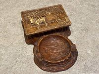 Closed view of ashtray and cigarette case with embossed details on lid showing horse-drawn carriage and inscriptions