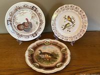 Three collectible plates displayed on stands showing bird and turkey themes and ornate rims on wooden surface.