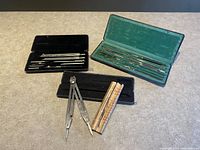 Three cases containing sets of metal drawing and drafting tools including compasses, dividers, and ruling pens, each case lined with fabric in black or green.